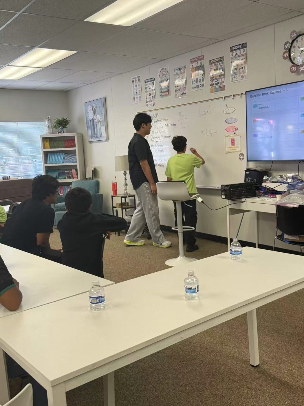 David Liu standing at the front of a classroom with children at long tables and a lesson on the screen behind him.