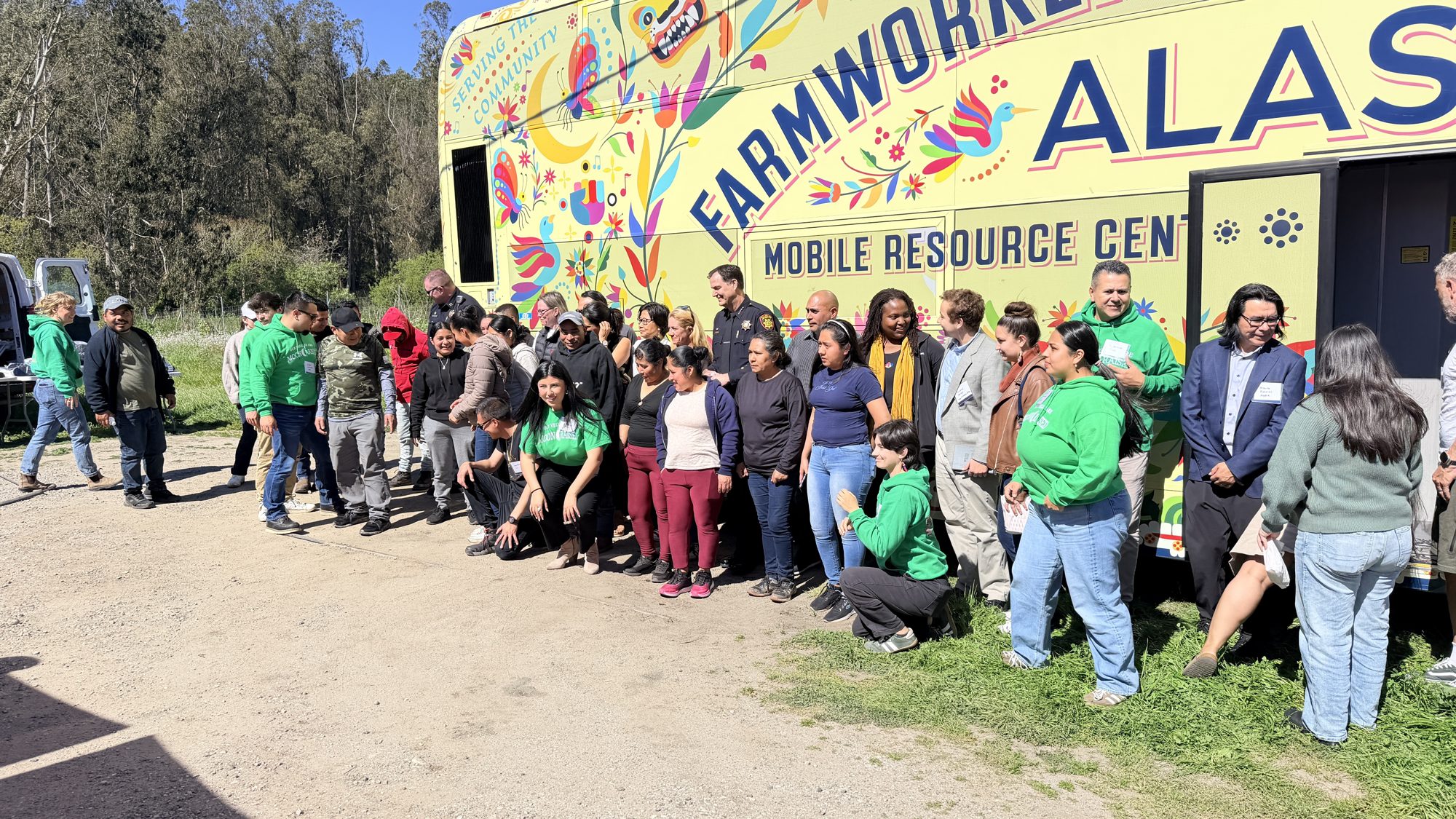 Volunteers and farmworkers gathered in front of the ALAS Farmworker Equity Express bus, a painted mobile resource center, on a farm.