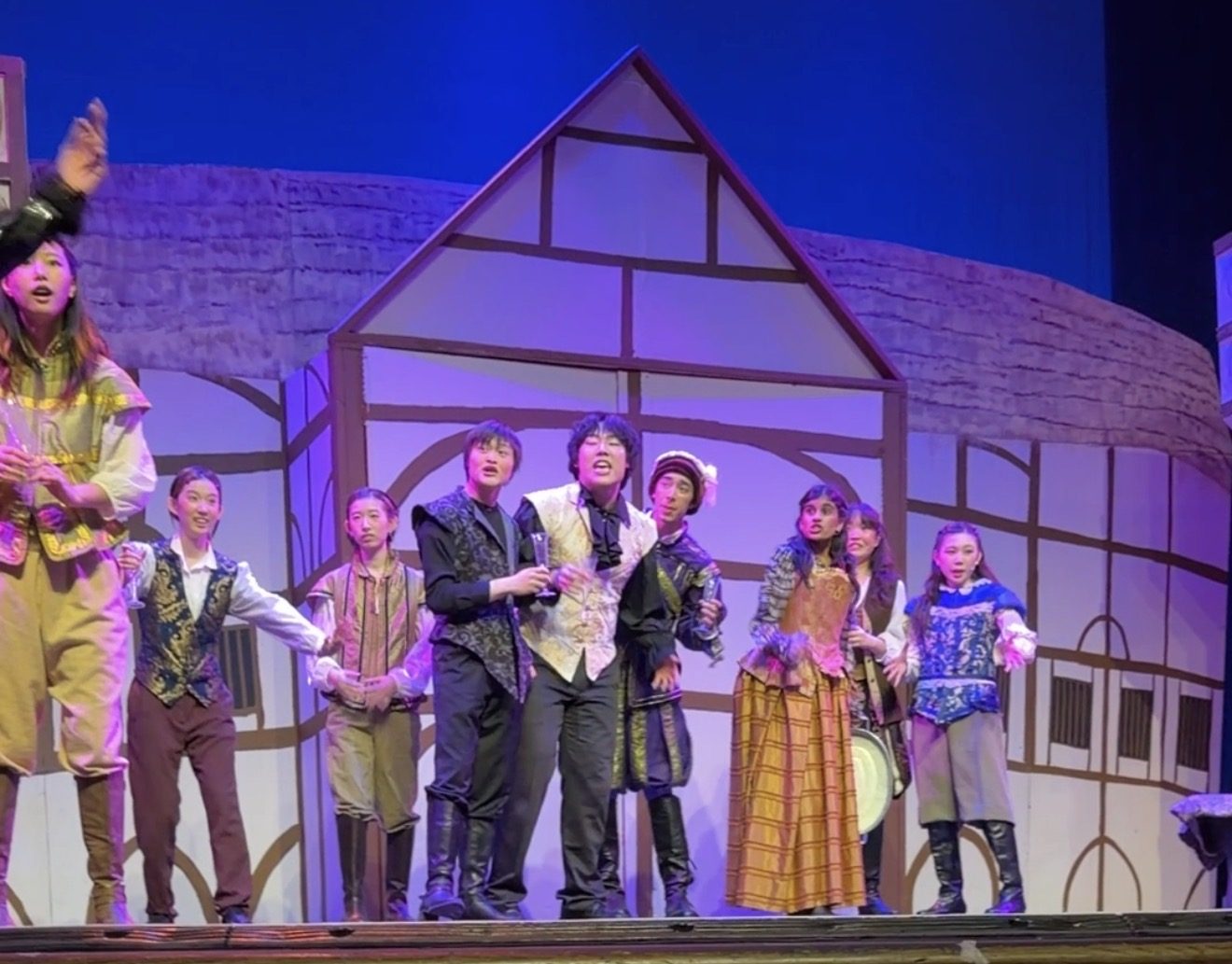 The cast of a school musical on stage in Renaissance costume in front of a painted half-timbered house.