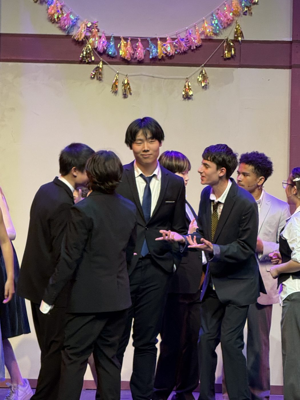 Students in suits gathered under strings of decorations backstage before a performance.