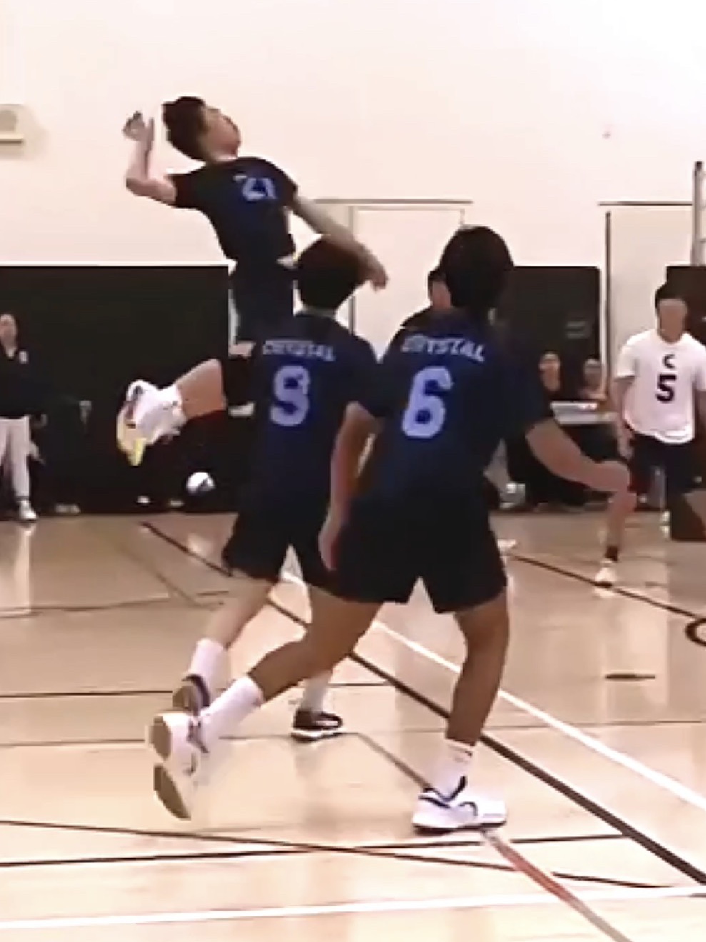A Crystal Springs Uplands player in a navy jersey, number 21, rising above two teammates to attack a set in a school gym.
