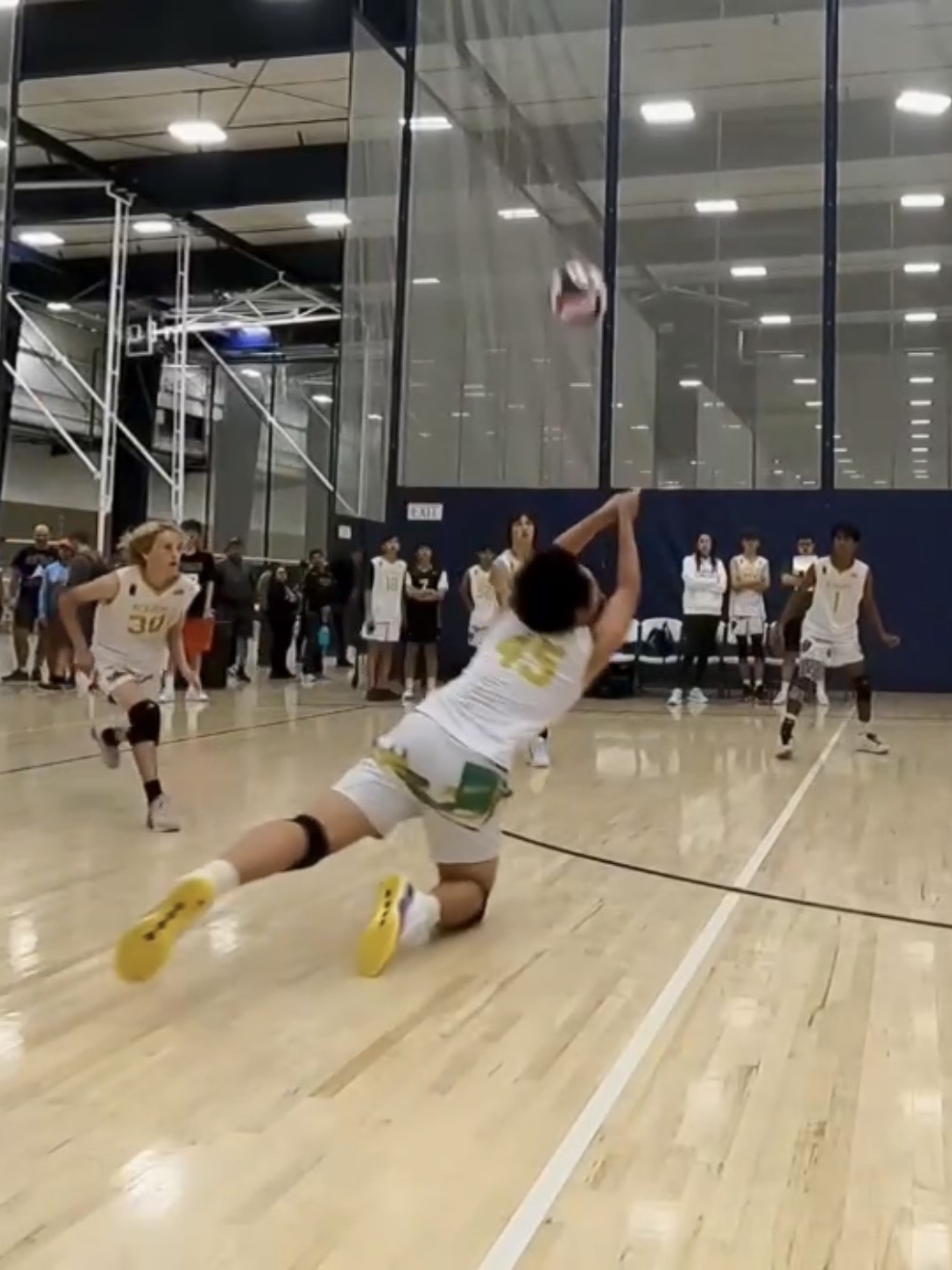 A club player in a white and green jersey, number 45, sliding to the floor to dig a ball in a multi-court facility.